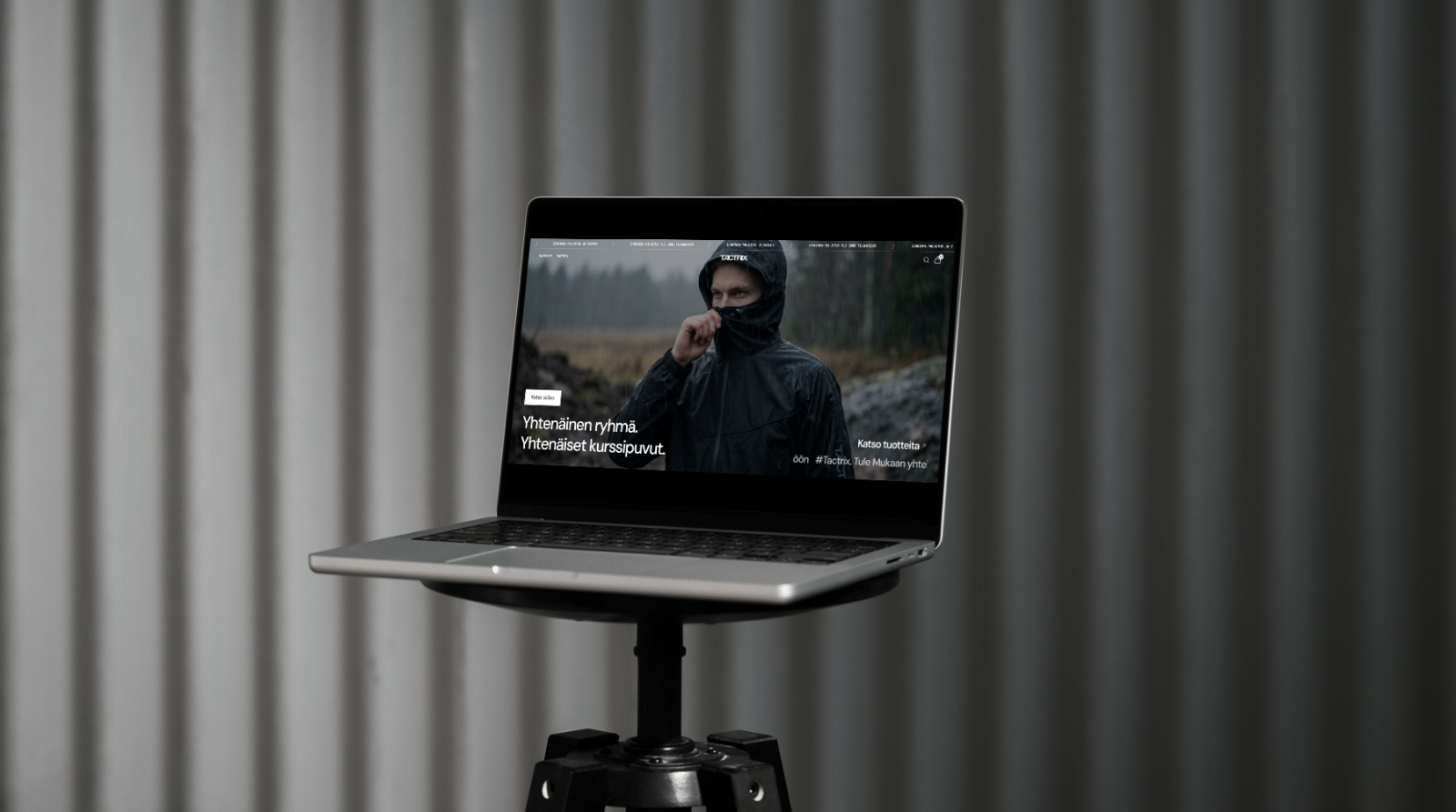 Tactrix outdoor clothing website displayed on a laptop on a black studio stand in front of grey curtains