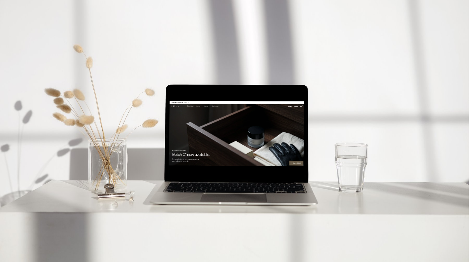 Ljóma skincare website displayed on a laptop on a bright white table beside dried stems and a glass of water